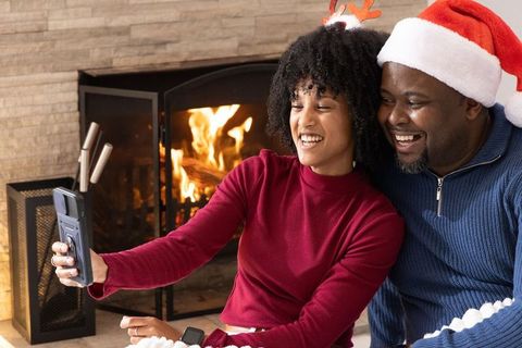 African american couple taking festive selfie by fireplace wearing santa hat and antlers