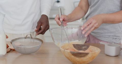 Multiracial couple whisking batter on wooden countertop, glass mixing bowl, eggshells
