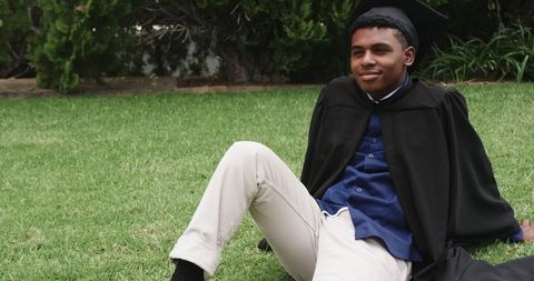 Relaxed african american graduate wearing gown and mortarboard sitting on campus lawn