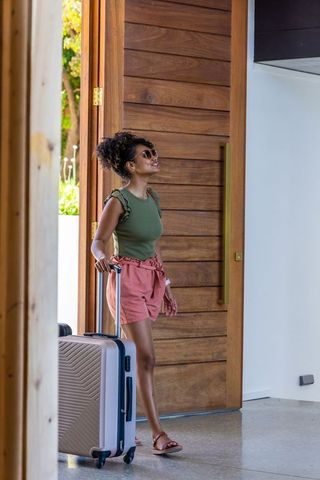 Woman standing at modern wooden pivot door pulling hard-shell suitcase wearing sunglasses