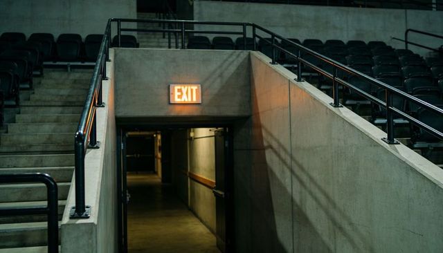 Glowing exit sign casting warm light over concrete arena stairwell and dim corridor