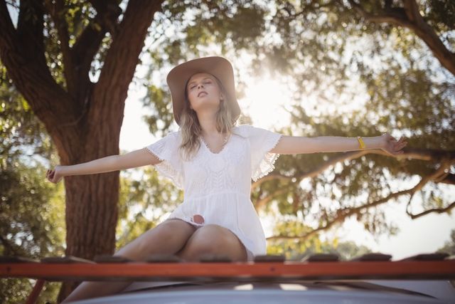 Young woman with arms outstretched sitting on van against tree