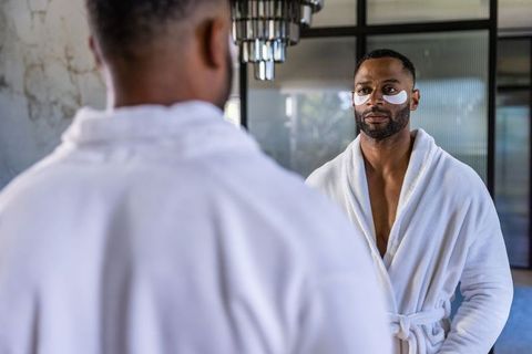 Mature african american man wearing eye patches and white robe practicing skincare routine