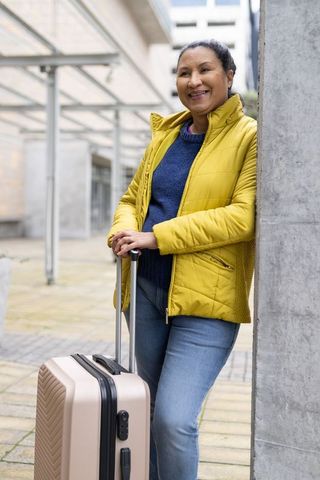 Smiling traveler leaning on column with suitcase in modern city plaza wearing yellow jacket