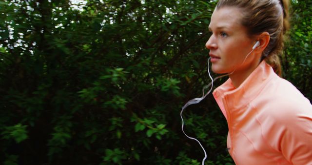 A young Caucasian woman is jogging in a lush green environment, focused and wearing earphones. Her active lifestyle and commitment to fitness are evident as she maintains her pace surrounded by nature.