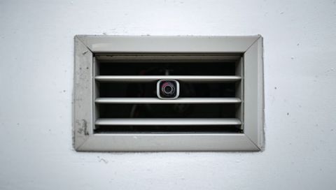 Square surveillance camera peeking through metal vent grille on scuffed white wall
