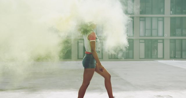 Young biracial woman walks through colorful smoke outdoors. She exudes confidence and style in a casual urban setting.