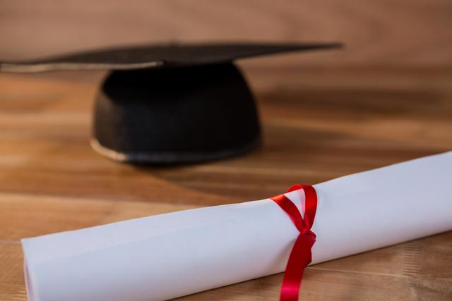 Close-up of a graduation certificate tied with a red ribbon and a mortar board on a wooden table. Ideal for use in educational materials, graduation announcements, academic achievements, and celebratory content.