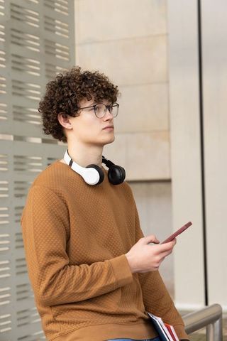 Teen studying outdoors wearing headphones, holding smartphone and folder by modern facade