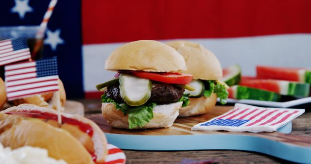 Hamburgers with American flag toothpicks are set against a backdrop of the United States flag, evoking a patriotic theme. The scene suggests a festive celebration, for a national holiday like the Fourth of July.