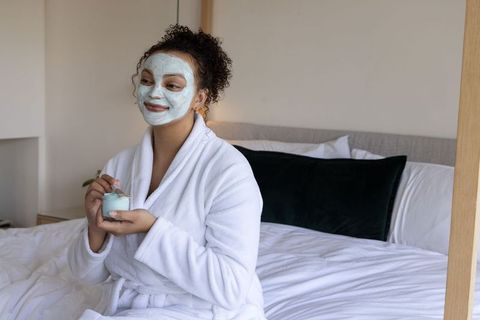 African american woman in robe applying facial mask on bed for at-home skincare relaxation