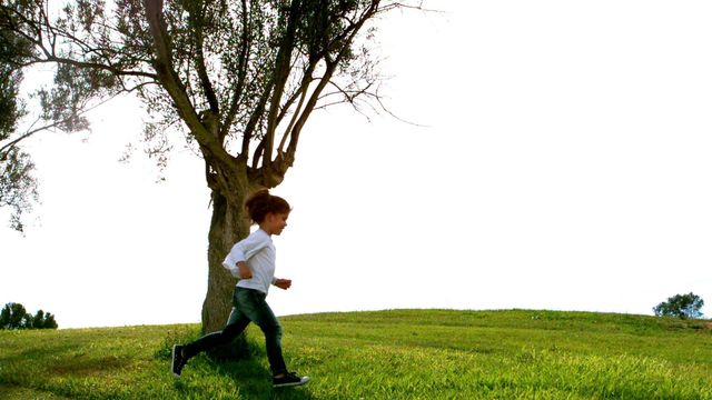 A young child is running energetically beside a tree in a grassy area during a bright day. This scene captures the joy and freedom of childhood play. Ideal for illustrating concepts of refreshing outdoor activity, promoting children's health or exploring nature. Suitable for use in advertisements, childhood development articles, and family-oriented content.