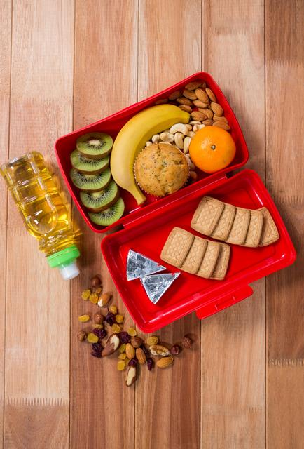 This image shows a healthy lunchbox containing a variety of fruits, nuts, and snacks, arranged on a wooden table. The lunchbox includes kiwi slices, a banana, an orange, a muffin, biscuits, and a small bottle of oil. This image is perfect for promoting healthy eating habits, balanced diets, and nutritious meal planning. It can be used in articles, blogs, or advertisements related to school lunches, healthy lifestyle choices, and meal preparation.