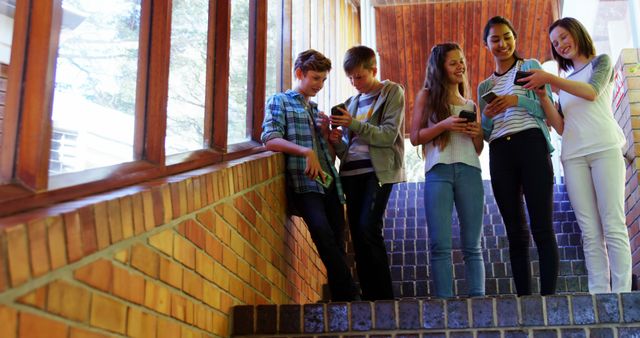 Diverse group of teenagers standing on stairway, engaging in social media on smartphones, laughing and interacting. Suitable for themes like teenage friendships, technology in youth, school environments, modern communication, and social media impact.