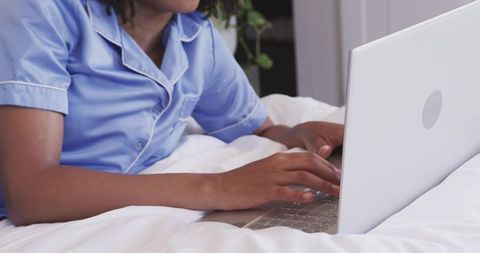 Typing on laptop in light blue pajamas on white bedding for remote work and cozy home