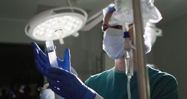 Healthcare professional wearing protective equipment holds and inspects syringe, ensuring its readiness for surgical procedure in a hospital operating room. Could be used for medical brochures, healthcare advertisements, educational materials about safety and health protocols, or professional healthcare websites.