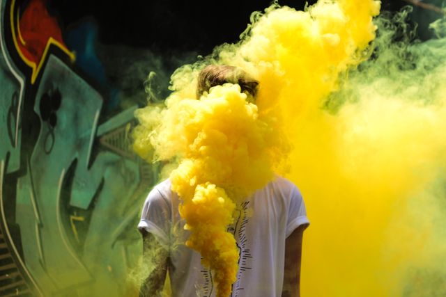 Person standing in an alleyway covered in graffiti, obscured by a dense yellow smoke bomb. Image highlights urban culture, creativity, and youth expression. Ideal for use in artistic projects, urban lifestyle blogs, and campaigns promoting street art and youthful energy.