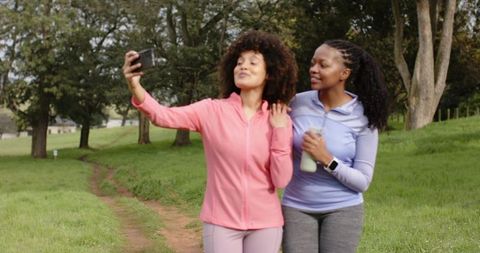 Diverse women walking park trail taking selfie in activewear holding water bottle