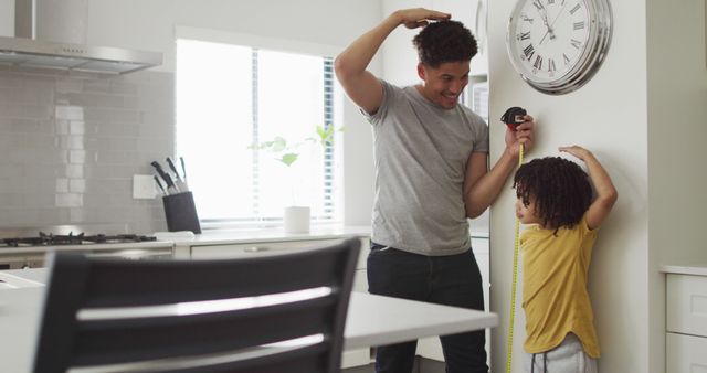Father using measuring tape to track son's height in modern kitchen, sunlight coming through the window. This image is great for promoting family togetherness, parenting, home life, childhood milestones, growth tracking, and family memories.