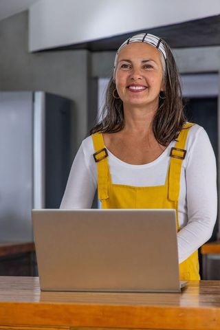 Middle-aged woman wearing yellow overalls using laptop in modern cozy kitchen