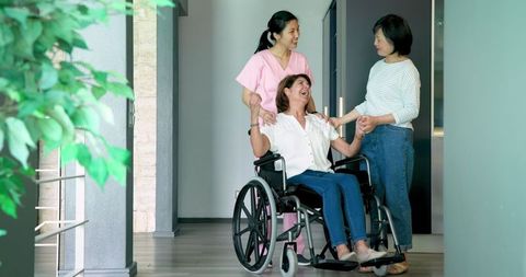 Elderly Woman in Wheelchair with Nurse and Friend in Care Facility Hallway