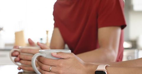 Diverse couple holding ceramic mugs at kitchen counter showing smartwatch and wedding ring