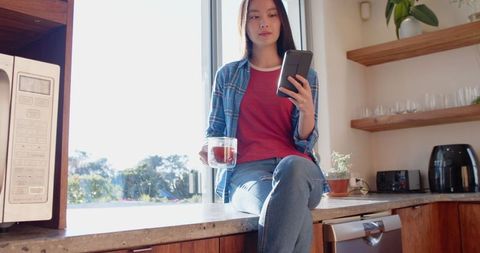 Young Woman Relaxing in Modern Kitchen with Smartphone and Mug