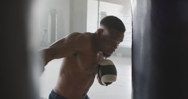 Depicting a professional boxer engaged in an intense training session with a punching bag in a gym setting. This can be used for fitness and health-related content, sports training blogs, promotional materials for gyms or martial arts studios, and motivational pieces that emphasize strength and dedication. Ideal for illustrating themes of physical fitness, determination, and athleticism.