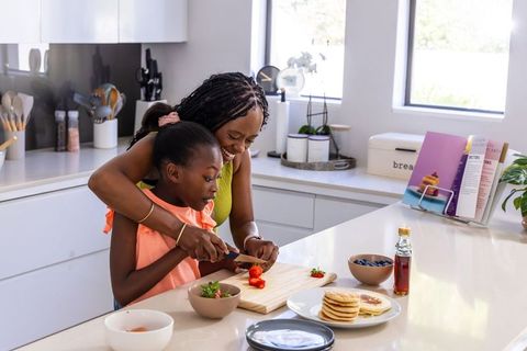 African mother guiding daughter slicing strawberries on kitchen counter with pancakes