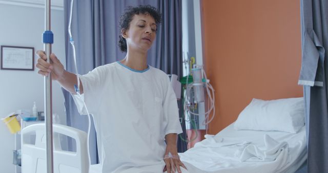 Female patient sitting on hospital bed, holding IV drip pole in hospital room. Ideal for healthcare and patient recovery, hospital environment visuals, medical treatment themes, and articles on patient care.