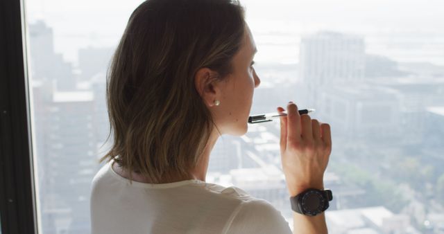 A woman is thoughtfully looking out a large office window while holding a pen against her lips, lost in consideration. The city's buildings and streets are blurred in the background. This image is perfect for use in business materials, websites, blog posts, or presentations to convey themes of contemplation, innovation, office environments, and professional work.