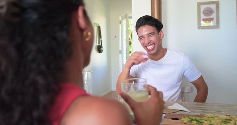 Young man laughing with friend over casual home meal, sharing drink and warm conversation