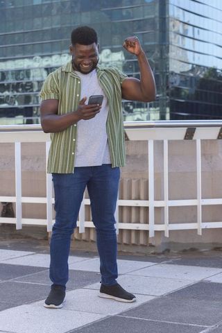 African man celebrating while checking smartphone on urban rooftop terrace with glass reflections