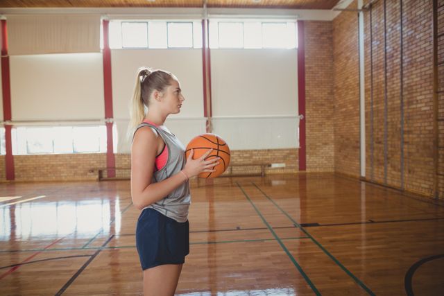High school girl standing in a gymnasium holding a basketball, looking determined. Ideal for use in educational materials, sports training guides, youth fitness programs, and motivational content related to sports and physical education.