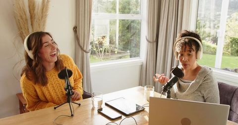 Two women recording lively home podcast with headphones, microphones and laptop