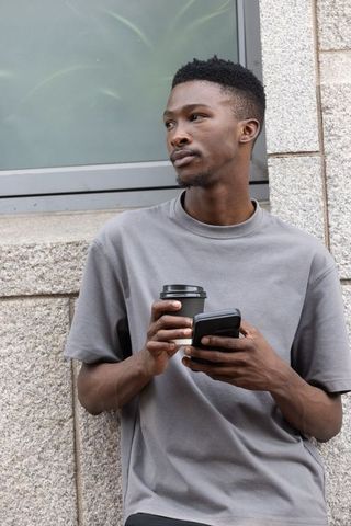 African man leaning on urban stone wall holding coffee and smartphone, street portrait