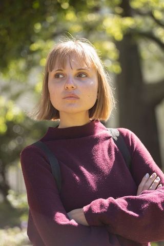 Young woman standing with arms crossed in sunlit park wearing burgundy sweater