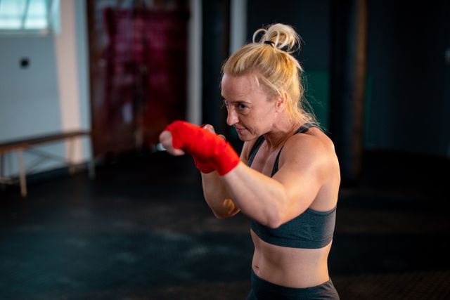 Caucasian woman in gym practicing shadow boxing with red hand wraps. Ideal for fitness, strength training, and martial arts content. Useful for promoting health, exercise routines, and athletic determination.