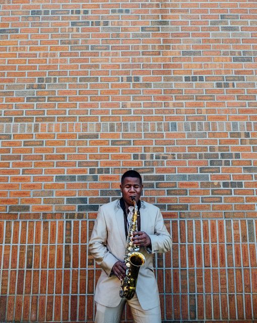 Man dressed in a formal suit playing saxophone against a textured brick wall. Can be used in promotions for music events, jazz festivals, educational materials on music, and street performance themes.