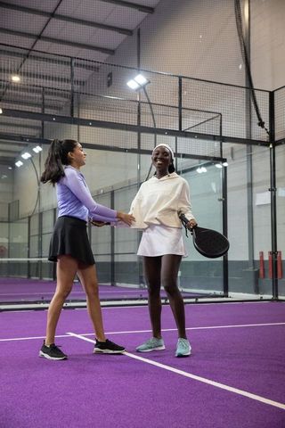 Female padel players smiling and shaking hands on purple court holding black racket