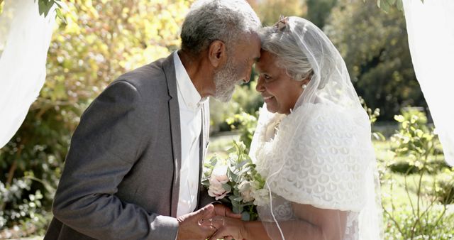 Happy diverse senior bride and groom holding hands and touching heads on wedding day in sunny garden. Summer, marriage, romance, love, celebration, tradition and senior lifestyle, unaltered.