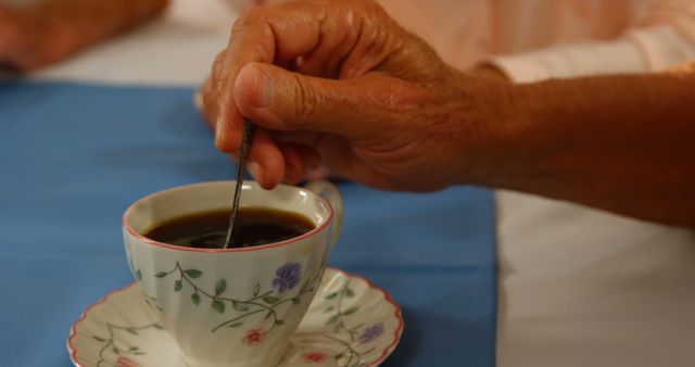This image captures an elderly hand stirring coffee in a beautifully floral teacup, resting on a matching saucer against a blue tablecloth backdrop. Ideal for use in articles related to senior lifestyle, casual moments, fine dining, coffee breaks, or comforting and relaxing scenes.