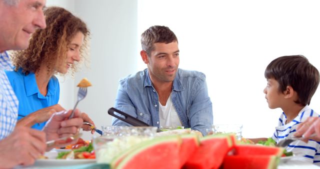 Family smiling and engaging in conversation while enjoying a fresh summer meal at home. A variety of colorful food is being shared, portraying a sense of togetherness and warmth. Perfect for illustrating family gatherings, healthy lifestyle, and summer dining.