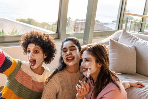 Diverse friends applying face masks taking selfie on sunlit living room couch