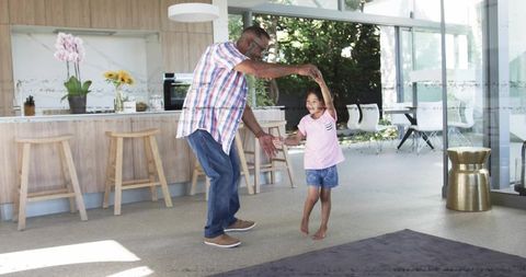 Man dancing with girl twirling barefoot beside kitchen island, sharing joyful family moment