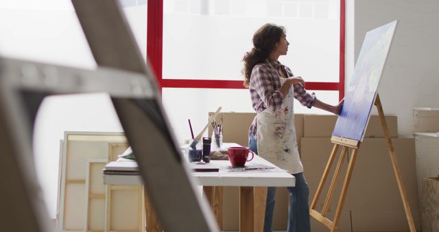 Female artist working on a canvas in a bright, well-lit studio. Ideal for use in articles or ads about creativity, art classes, hobbies, or feminine artistic expression. Can also be used to depict home art spaces or painting tutorials.