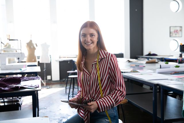 Young red-haired Caucasian female fashion student smiling in a design studio, holding a tablet with a yellow tape measure around her neck. Ideal for use in educational materials, fashion industry promotions, creative workspace advertisements, and articles about fashion design education.