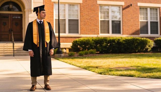 Graduate wearing black gown and gold stole standing on campus walkway celebrating success