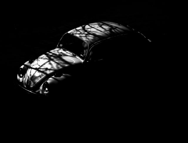 Vintage car partially in shadows with tree branches reflecting on the surface. Classic and retro look with dramatic use of light and darkness. Suitable for art prints, automotive advertisements, and moody artistic works.