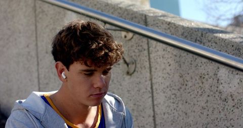 Young man sitting on concrete steps listening with earbuds wearing hoodie, urban contemplative